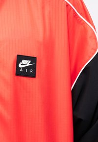 Chaqueta Nike Air en naranja brillante y negro, con un parche de logo negro y ribetes blancos a lo largo de la costura del hombro.