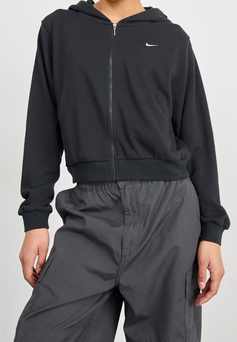 En person iklädd en svart zip-up hoodie med en liten vit Nike-logotyp, kombinerad med löst sittande mörkgrå cargobyxor mot en plain bakgrund.