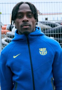 Μπλε ζακέτα με φερμουάρ από μαλακό ύφασμα, με κίτρινο λογότυπο Nike και σήμα της Μπαρτσελόνα. Ορατές ραφές και σχέδιο ψηλού γιακά.