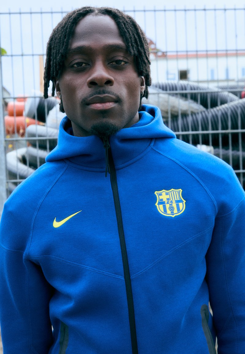 Μπλε ζακέτα με φερμουάρ από μαλακό ύφασμα, με κίτρινο λογότυπο Nike και σήμα της Μπαρτσελόνα. Ορατές ραφές και σχέδιο ψηλού γιακά.