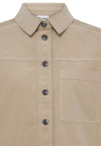 Chemise en velours côtelé beige avec col pointu, cinq boutons métalliques sur le devant et une poche plaquée sur la poitrine gauche.