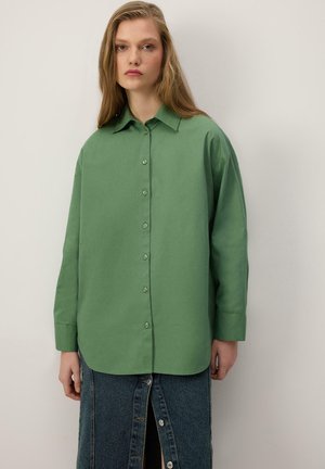 Groen overhemd met lange mouwen en knopen, gemaakt van gladde stof. Heeft een ontspannen pasvorm en bijpassende knopen. Draagt bij een denim rok.