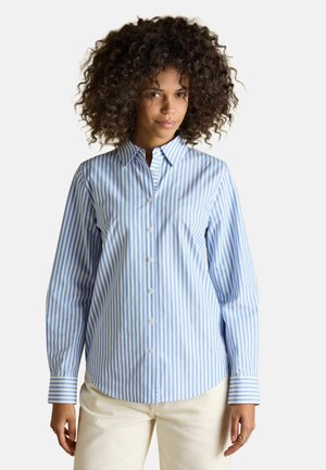 Woman with curly hair wearing a blue and white vertical striped button-up shirt and light-colored pants, standing against a plain background.