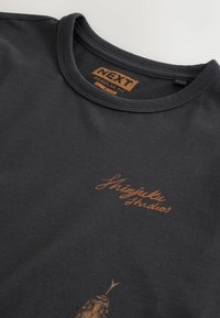 T-shirt noir à col rond avec texte orange « Shinjuku Studios » et petit graphique de poisson, étiquette "NEXT Regular Fit M" à l'intérieur du col.