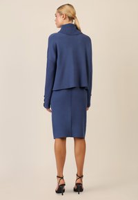 Maglione blu navy con colletto alto e spalle scese, abbinato a una gonna a tubino blu navy aderente. Entrambi i capi presentano una texture liscia.