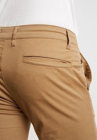 Pantaloni beige realizzati in un tessuto morbido con una superficie liscia. Presentano una tasca posteriore e cuciture sopraffinate, con un girovita a media altezza.