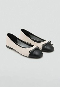 Zapatos planos beige con punteras redondeadas negras y pequeños lazos decorativos sobre un fondo gris claro.