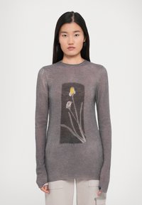 Femeie îmbrăcată cu un pulover gri ajustat cu un imprimeu floral rectangular, caracterizat printr-o floare galbenă și albă, asortat cu pantaloni de culoare deschisă.