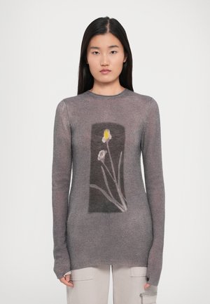 Donna che indossa un maglione grigio aderente con una stampa floreale rettangolare che presenta un fiore giallo e bianco, abbinato a pantaloni chiari.
