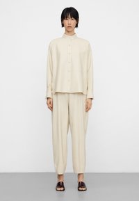 Beige button-up shirt with a chest pocket, paired with loose-fitting beige pants and dark brown slip-on sandals, set against a plain background.