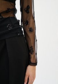 Frau in einem durchsichtigen, schwarzen Mesh-Oberteil mit langen Ärmeln, verziert mit schwarzen Pailletten in Blumenform, und hochgeschnittenen schwarzen Hosen, Arm entspannt an der Seite.