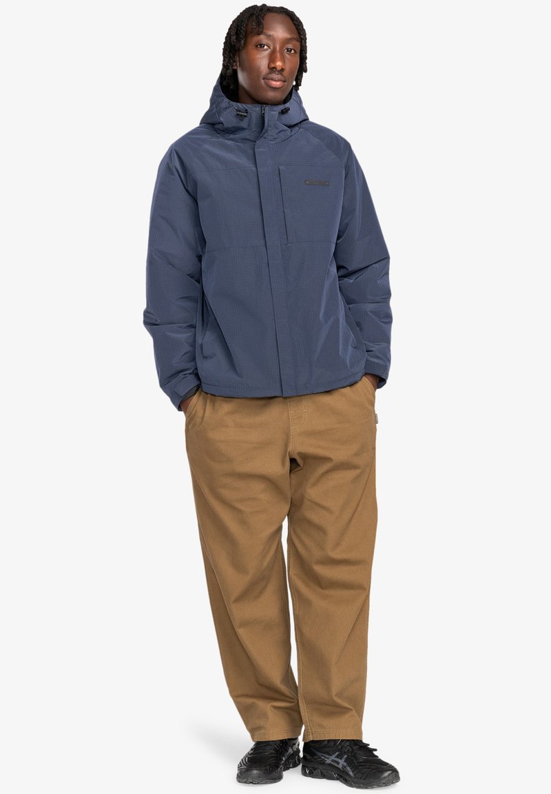 Marineblaue isolierte Jacke mit Kapuze, ausgestattet mit einem Reißverschluss vorne und einer Brusttasche. Kombiniert mit khakifarbenen Hosen und schwarzen Schuhen.