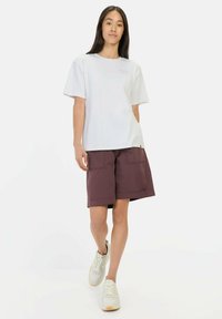 Weißes T-Shirt mit kurzen Ärmeln und einem kleinen lila Grafik, kombiniert mit tiefburgunderfarbigen Cargo-Shorts und weißen Sneakers. Das Outfit ist lässig und entspannt.