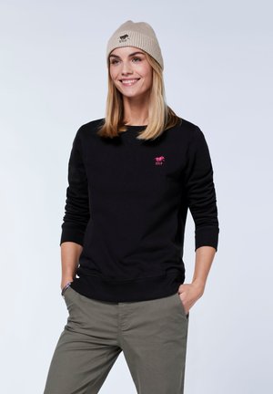 Black sweatshirt with round neckline and ribbed cuffs, featuring a small pink logo. Paired with olive green trousers and a light beige knit beanie.