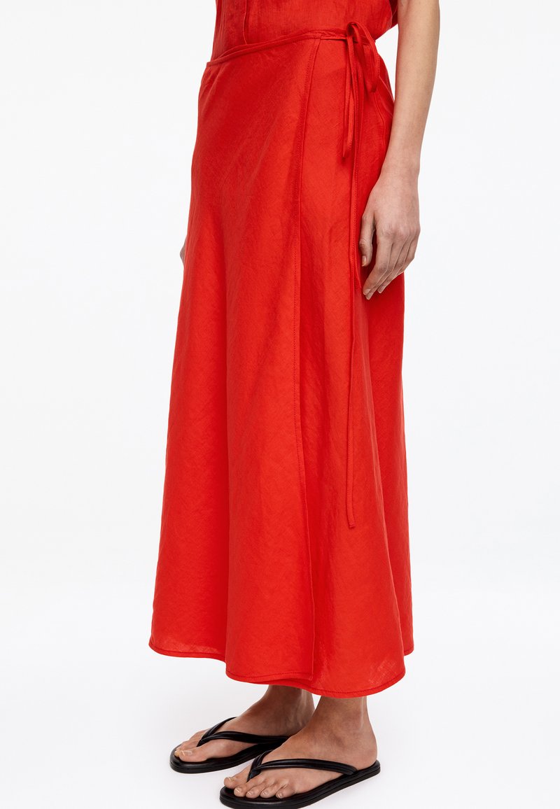Femme portant une jupe portefeuille rouge vif longueur cheville nouée à la taille, associée à des tongs noires simples, sur un fond blanc uni.