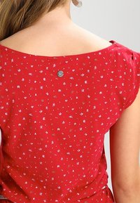 Naisen yläselkä punaisessa mekossa, jossa on pieniä valkoisia kukkakuvioita ja pyöreä metallinen emblem kaula-aukossa.