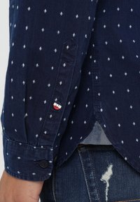 Tmavě modrá košile s bílou vyšívanou diamantovou výšivkou, manžetami na knoflíky a kontrastním knoflíkem. Měkká tkanina s mírným leskem a strukturovaným střihem.