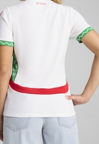 Persona vestida con una camiseta polo blanca con puños de mangas de patrón verde, paneles laterales verdes y una franja roja en la parte posterior, combinada con jeans de color azul claro.