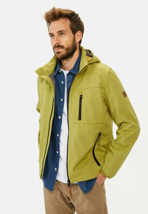 Lys grøn softshell-jakke med hætte, der har for- lommer og en lynlåslomme på brystet, båret over en blå skjorte og tanfarvede bukser.