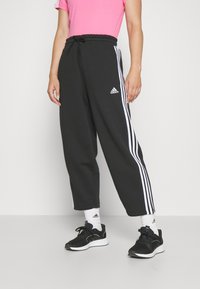 Svarta sweatpants med vita ränder längs sidorna, elastisk midja med dragsko, i kombination med svarta träningsskor och vita strumpor.