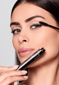 Czarna tuba tuszu do rzęs z napisem "Vamp! All in One", trzymana przez osobę z błyszczącymi ustami i warstwowymi rzęsami, na jasnoszarym tle.