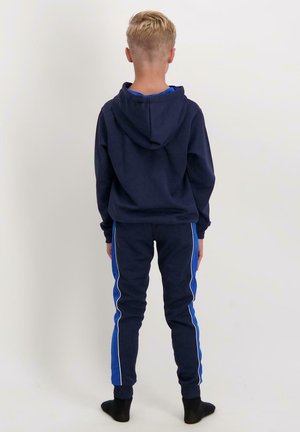 Jongen met kort blond haar staat met zijn rug naar de camera en draagt een marineblauwe hoodie en bijpassende sweatpants met blauwe zijstrepen en zwarte sokken.