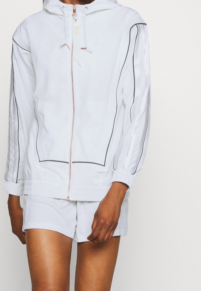 Vit atletisk jacka med huva, med svarta detaljer, en roséguld dragkedja och elastiska ärmslut. Kombinerad med matchande shorts.