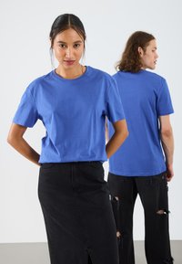 Tricou din bumbac albastru cu mâneci scurte, guler rotund, purtat de un model în față; blugi skinny negri și blugi uzati purtați de un alt model în spate.