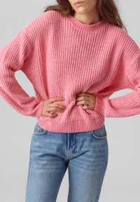 Rosa gestrickter Pullover mit einem strukturierten, gerippten Muster und einem runden Ausschnitt, kombiniert mit blauen Jeans. Der Schnitt ist lässig mit umgeschlagenen Ärmeln.