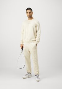 Krämfärgad sweatshirt med matchande mjukisbyxor, med strukturerad väv och elastiska manschetter, i kombination med en tennisracket och träningsskor.