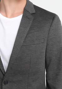 Primo piano di una persona che indossa un blazer grigio sopra una maglietta bianca a girocollo, con attenzione al torso superiore e ai dettagli dell'abbigliamento.