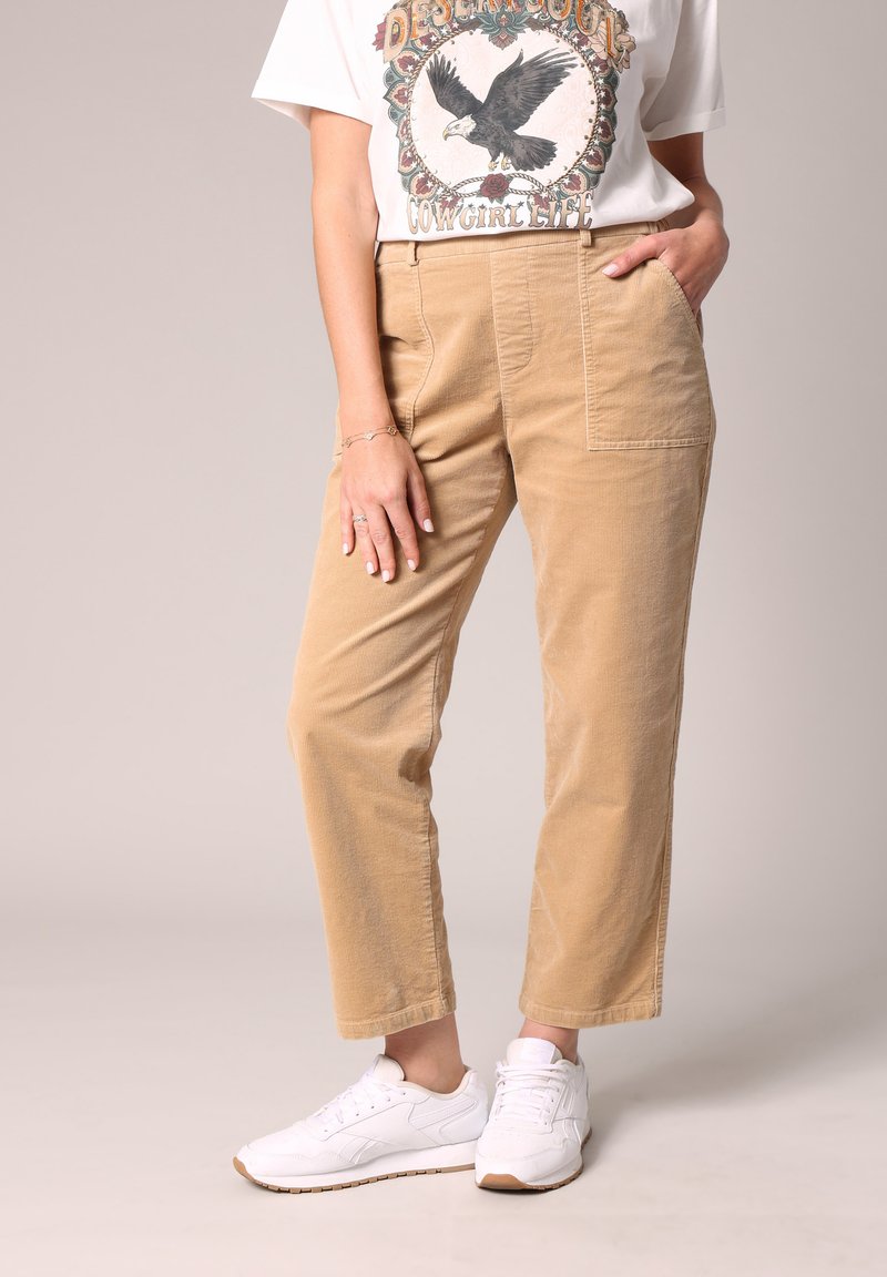 Pantalon en velours côtelé beige avec une coupe droite, doté de poches avant et d'une texture subtile, associé à des chaussures de sport blanches.