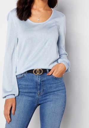 Mujer con suéter de manga larga azul claro, vaqueros azules, cinturón negro con hebilla doble en forma de anillo dorado y collar con colgante en forma de lágrima.