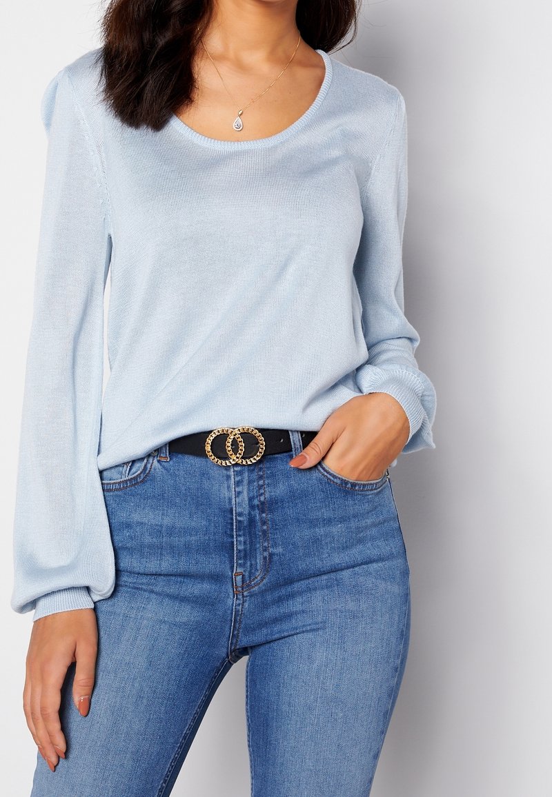 Femme portant un pull léger bleu clair à manches longues, un jean bleu, une ceinture noire avec une boucle double anneau dorée, et un collier pendentif en forme de goutte.