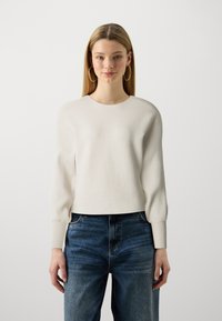 Light beige ribbed knit sweater with a round neckline and long, slightly puffed sleeves, paired with high-waisted blue jeans.