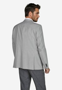 Helle graue maßgeschneiderte Sakkojacke mit einem Rückenschlitz, strukturiertem Stoff und drei schwarzen Knöpfen am Ärmel. Kombiniert mit grauen gemusterten Hosen.