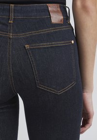 Jeans de mezclilla azul oscuro con costuras naranjas, etiqueta de cuero en la cintura y un bolsillo trasero con un detalle de botón metálico.