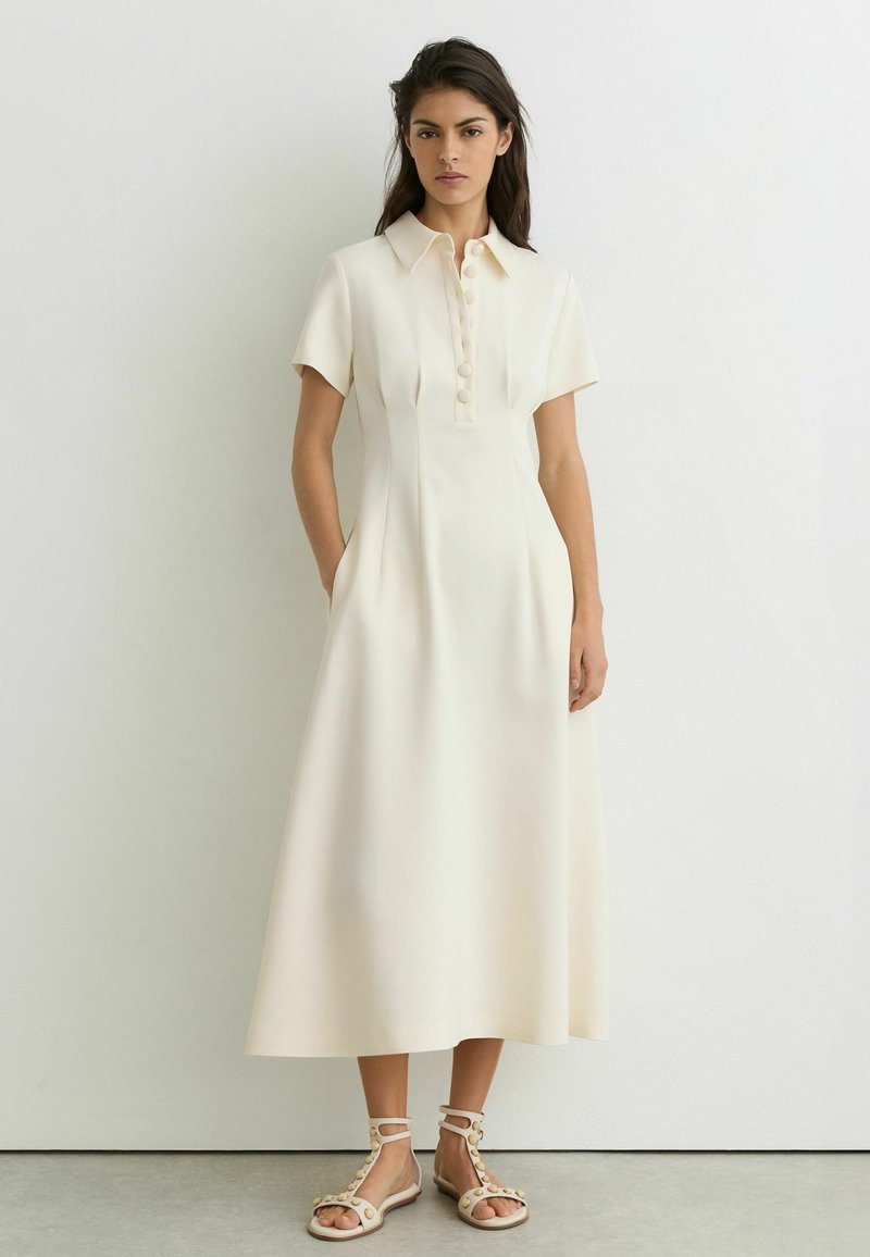 Jeune femme portant une longue robe crème à manches courtes avec des boutons sur le devant, debout contre un mur blanc uni.