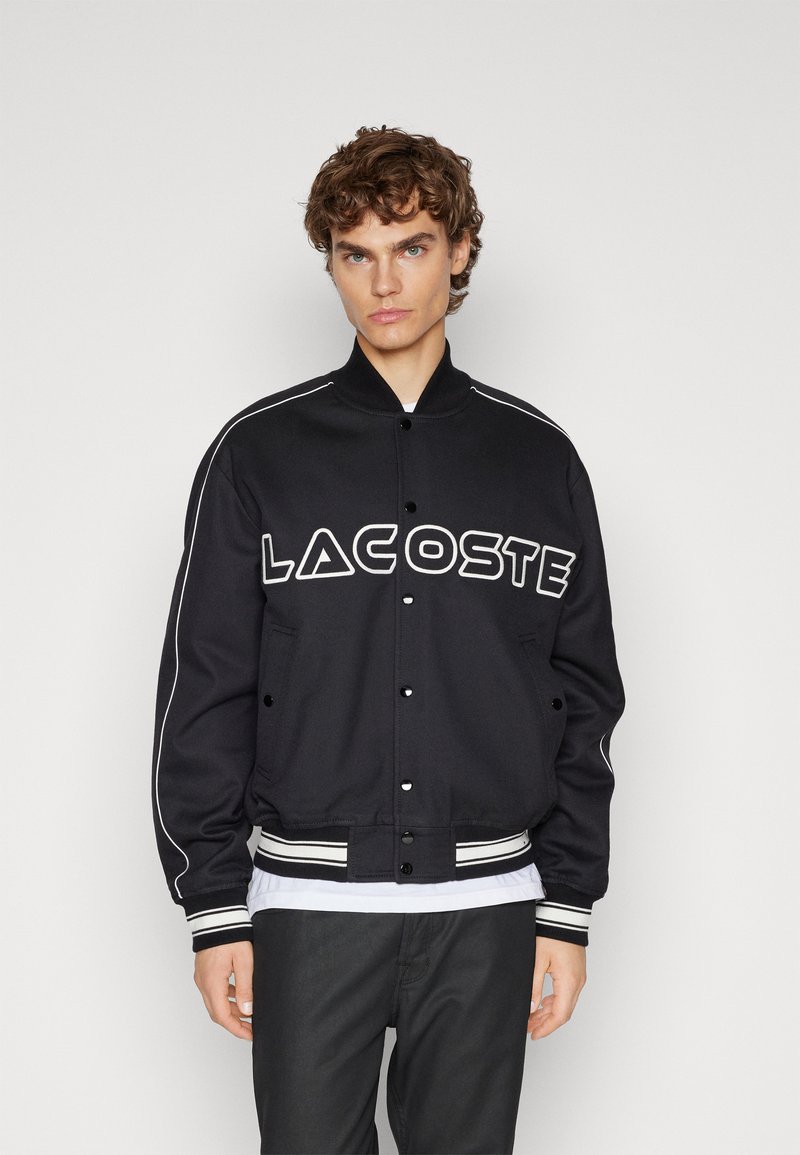 Veste varsity noire avec lettrage "LACOSTE" blanc, ourlet et poignets côtelés, et poches latérales. Présente des passepoils blancs le long des épaules.