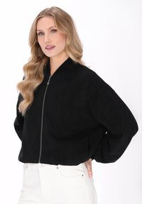 Chaqueta bomber negra con cierre de cremallera y acabado texturizado, con hombros caídos y puños acanalados. Combinada con pantalones blancos.
