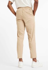 Beige, schmal zulaufende Hose mit elastischem Bund und Gesäßtasche. Glattes Material, kombiniert mit weißen Sneakers. Von hinten dargestellt.