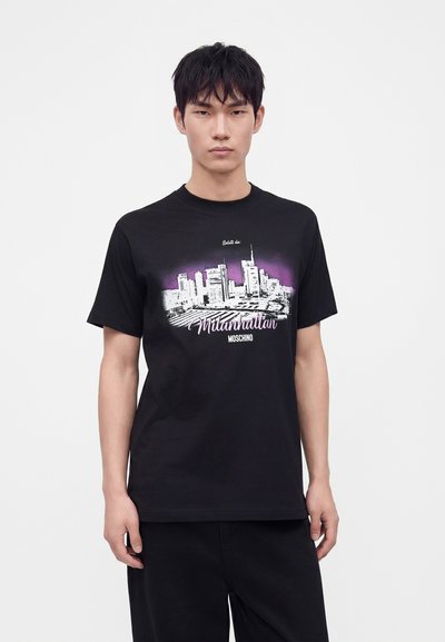 Jeune homme portant un t-shirt noir avec un graphisme de silhouette de la ville en blanc et violet et le texte "Milanhattan Moschino" sur le devant.