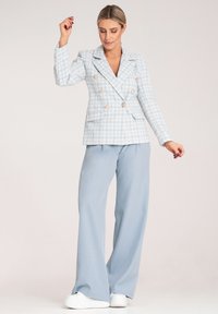 Jasnoniebieska podwójnie zapinana marynarka w kratę, z złotymi guzikami i kieszeniami, w połączeniu z jasnoniebieskimi spodniami o szerokich nogawkach i białymi sneakersami.