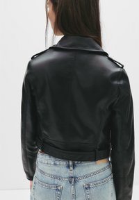 Chaqueta biker corta de cuero negro con cuello de solapa y hombreras. Cuenta con una cintura con cinturón y una textura suave. Combinada con jeans azul claro.