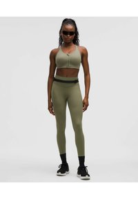 Olivengrünes Sportoutfit bestehend aus einem gekürzten Top mit einem Reißverschluss vorne und hochgeschnittenen Leggings. Strukturiertes Material mit glatter Oberfläche.