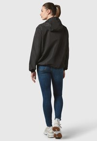 Black hooded jacket made of lightweight fabric, featuring a relaxed fit. Worn over blue jeans and light-colored sneakers.