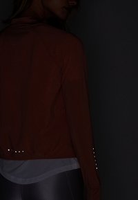 Persona que lleva una chaqueta roja oscura con puntos reflectantes en la espalda y las mangas, camiseta de malla gris debajo y leggings oscuros brillantes, contra un fondo negro.