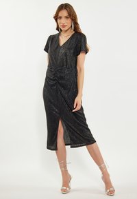 Rochie neagră, până la genunchi, cu decolteu în V, mâneci scurte, față plisată și o tăietură pe lateral. Material texturat cu luciu metalic; asortată cu tocuri argintii.
