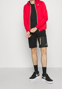 Červená zip-up bunda s kapucí, černé tričko, černé šortky s žlutým detailem zipu a černé tenisky Nike s bílou podešví.