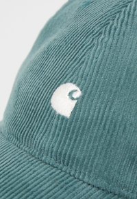 Blågrønn kordfløyelslue med en hvit brodert logo. Teksturyta med vertikale ribber og buet brem.